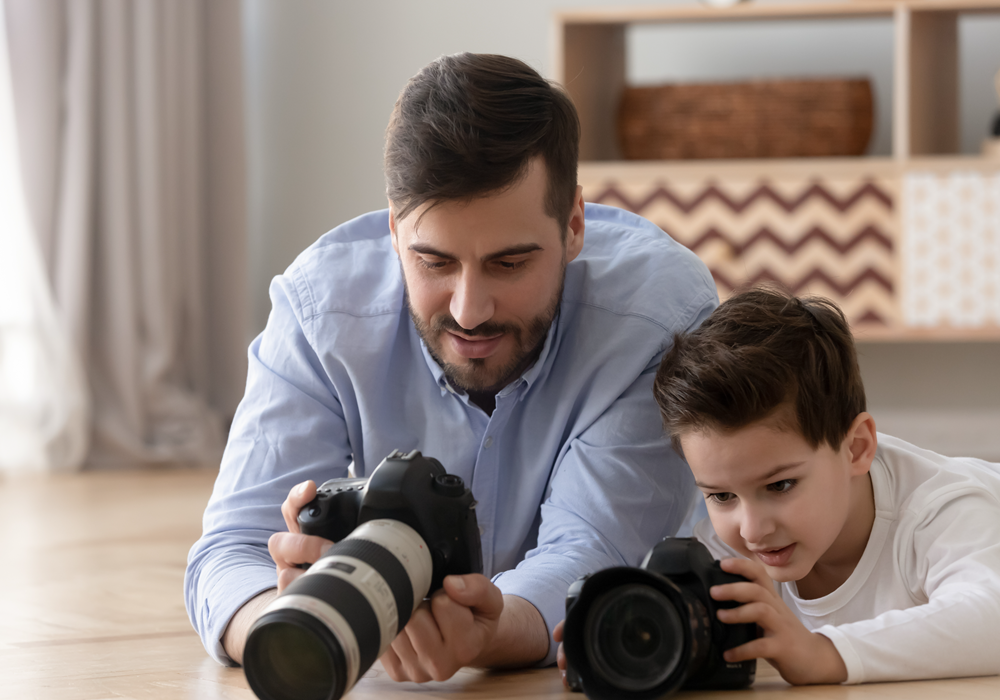  Father teaching son how to use a camera while laying on their apartment's hardwood-styled flooring.